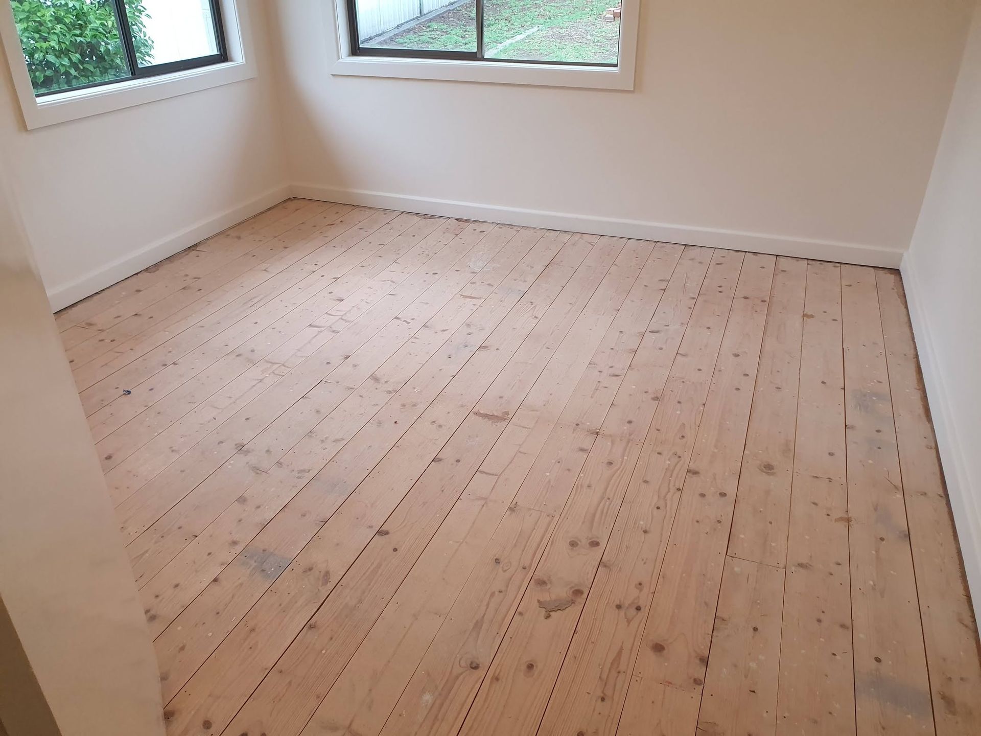 Empty room with unfinished wooden floor and two windows, white walls and baseboards