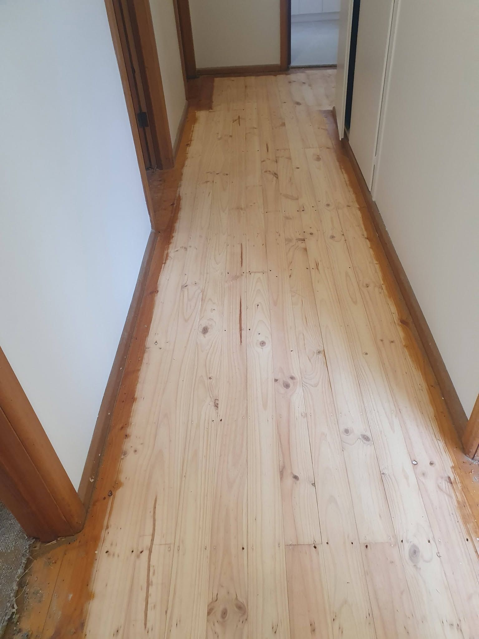 Narrow hallway with light wood floor, white walls, and wooden door frames leading to a room.