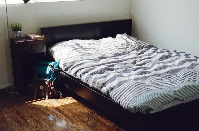 Bedroom with unmade bed, dark brown headboard, wooden floors, and a small nightstand.