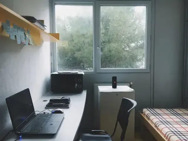 Desk with laptop near window, microwave, and small fridge in a simple room with a bed. Rain outside.