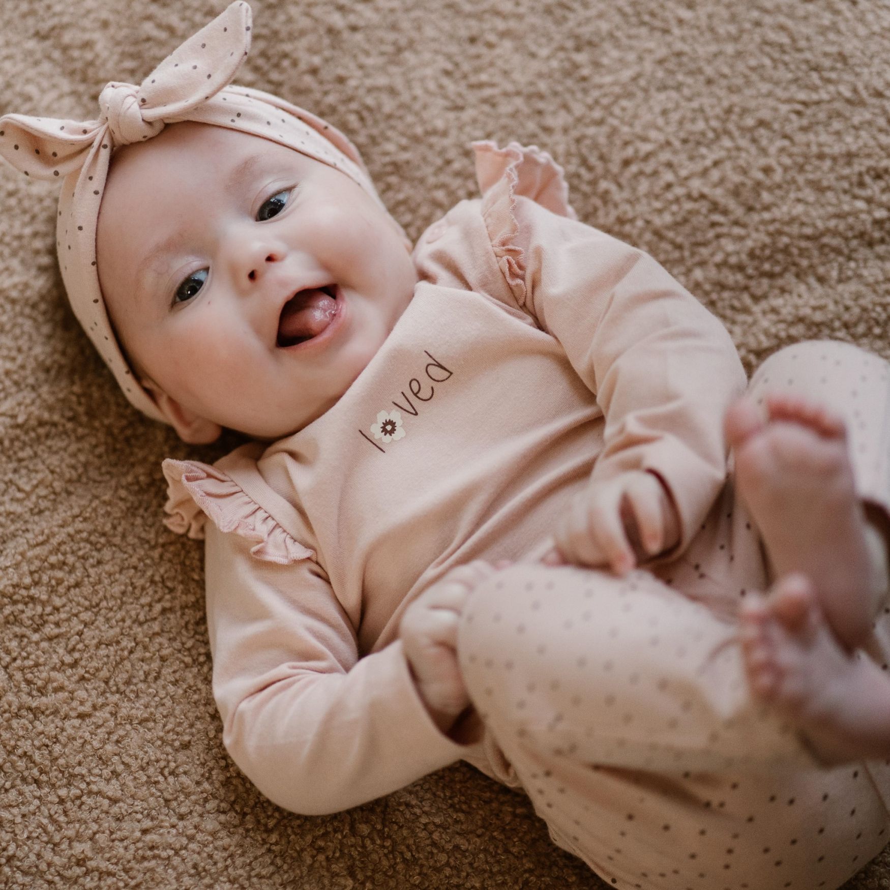 Een lachende baby in een beige outfit en met een haarbandje ligt op een tapijt met de voetjes omhoog.
