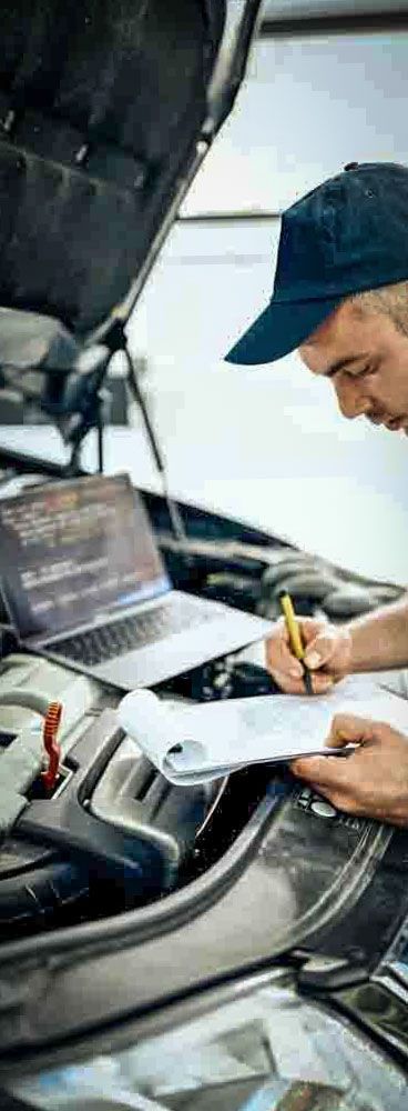 am auto mechanic writing up a report on a clipboard at the front of a vehicle with its hood up.