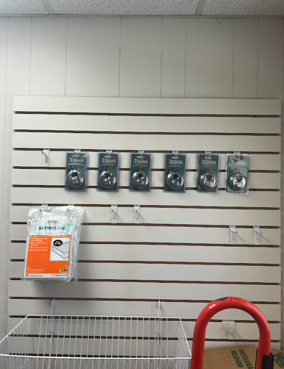 A white slatwall display holds six small, identical packaged locks in a row, with a hanging bag and a wire bin below.