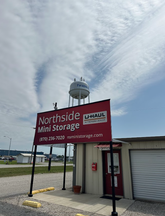 A red sign for Northside Mini Storage with a U-Haul logo sits under a water tower against a blue, partly cloudy sky.