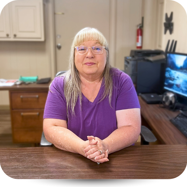 A person with blonde hair and glasses wearing a purple shirt sits at a desk with their hands clasped in an office.
