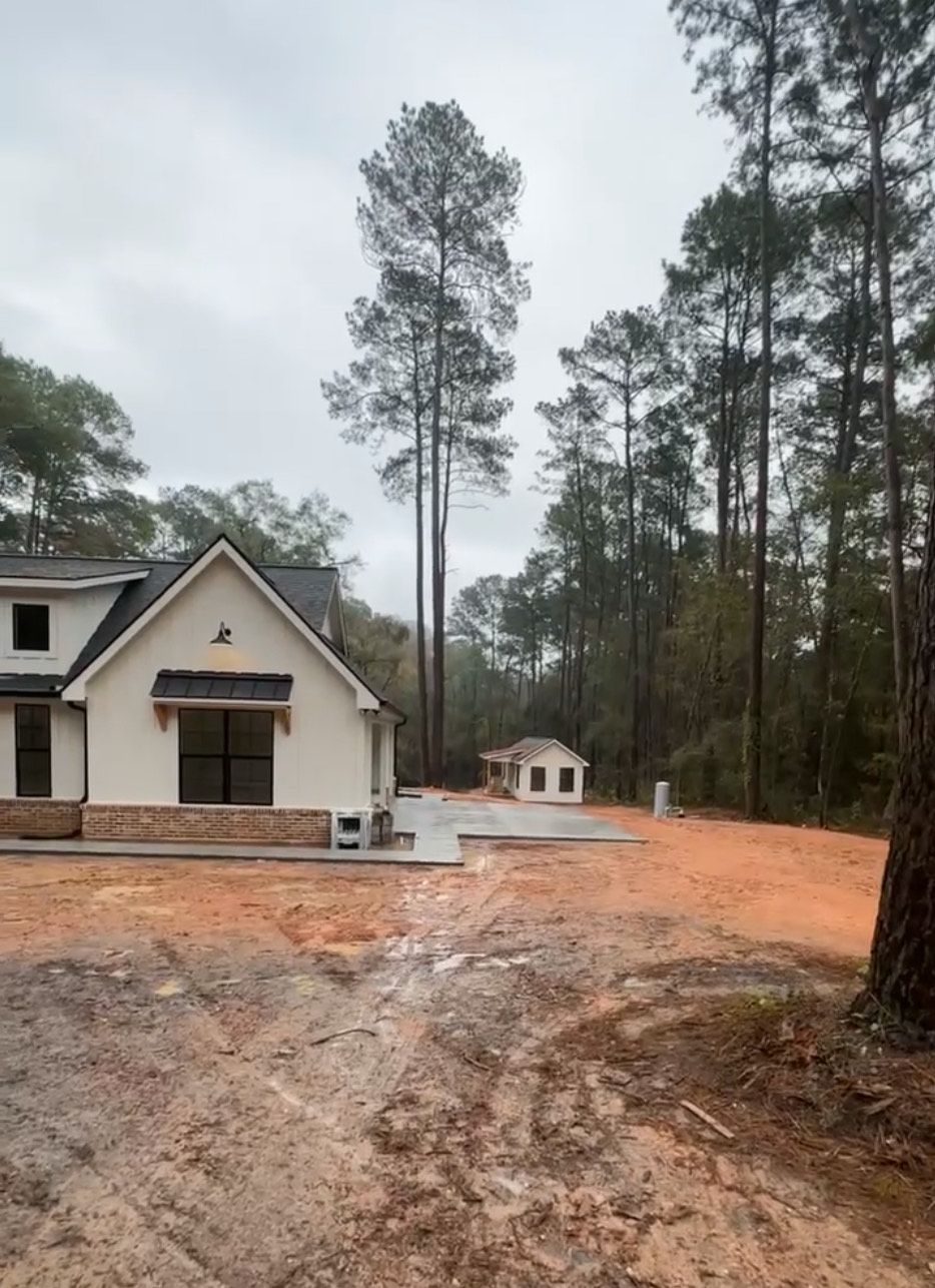New house under construction in a wooded area. A small building sits to the right of the house. The ground is dirt.