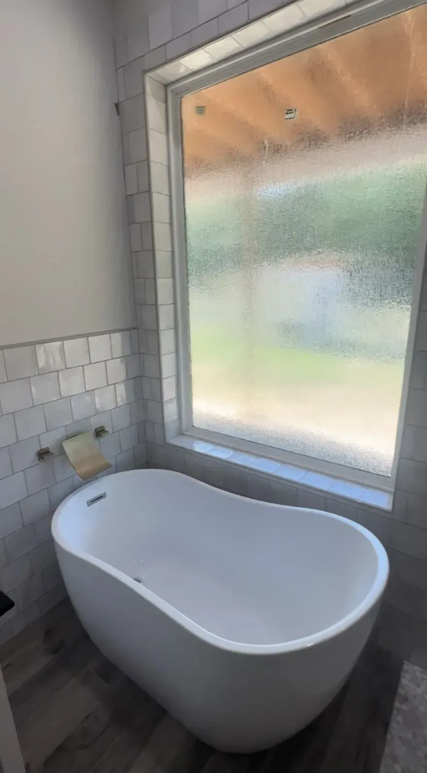 A white, oval-shaped bathtub sits beneath a window in a modern bathroom with gray tile and wood-look flooring.