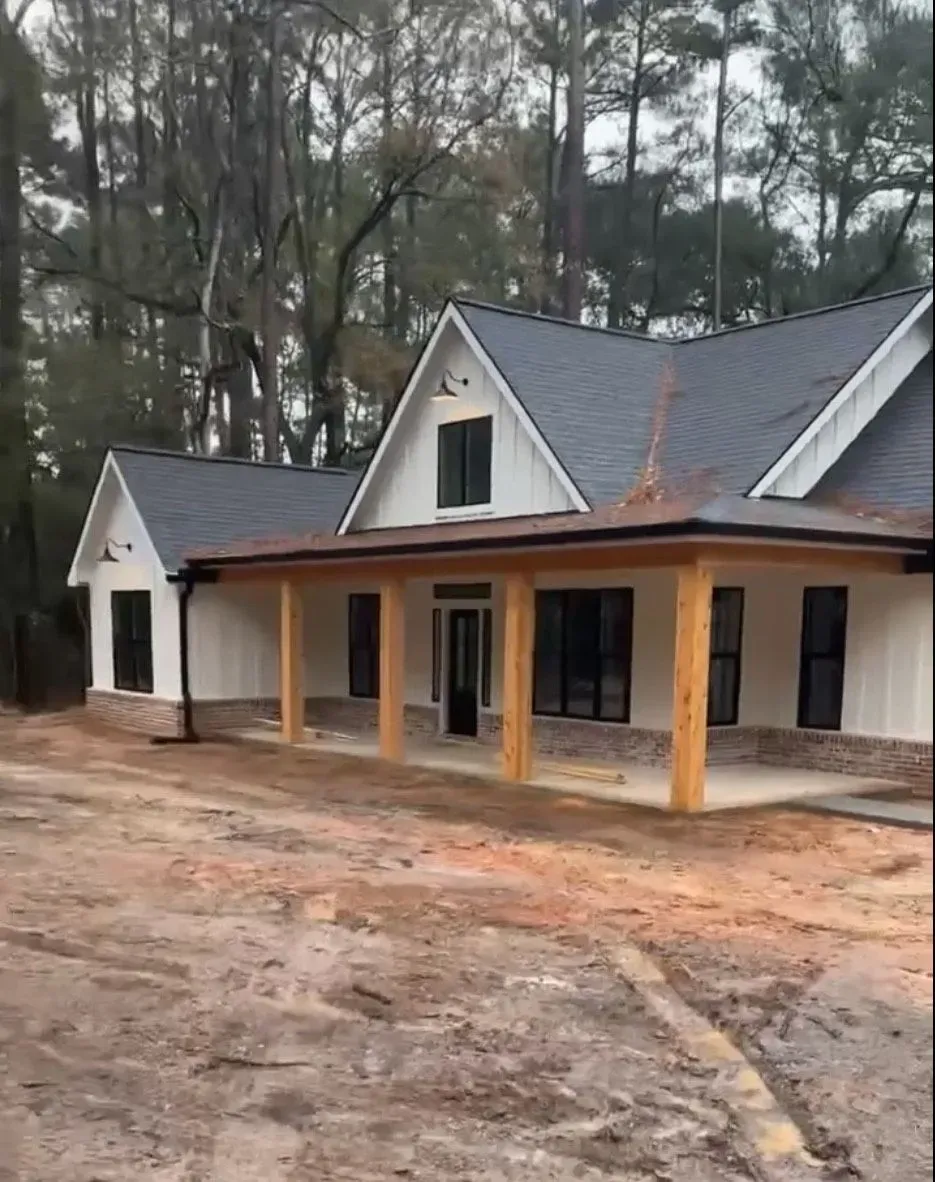 White farmhouse with a black roof and porch, set in a dirt yard with trees in the background.