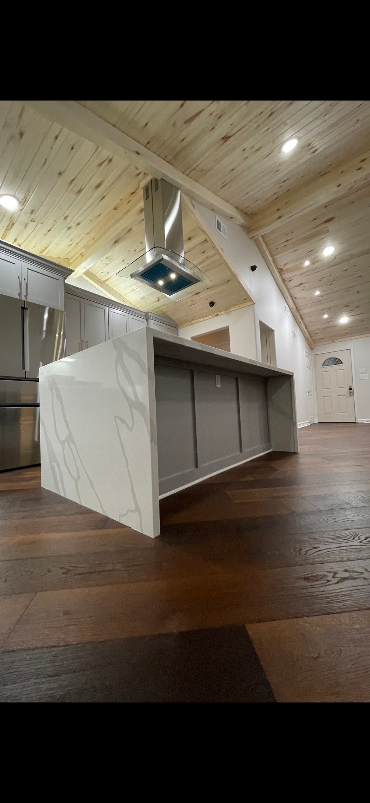 A modern kitchen with a large white countertop island, stainless steel appliances, and a wooden ceiling and floor.