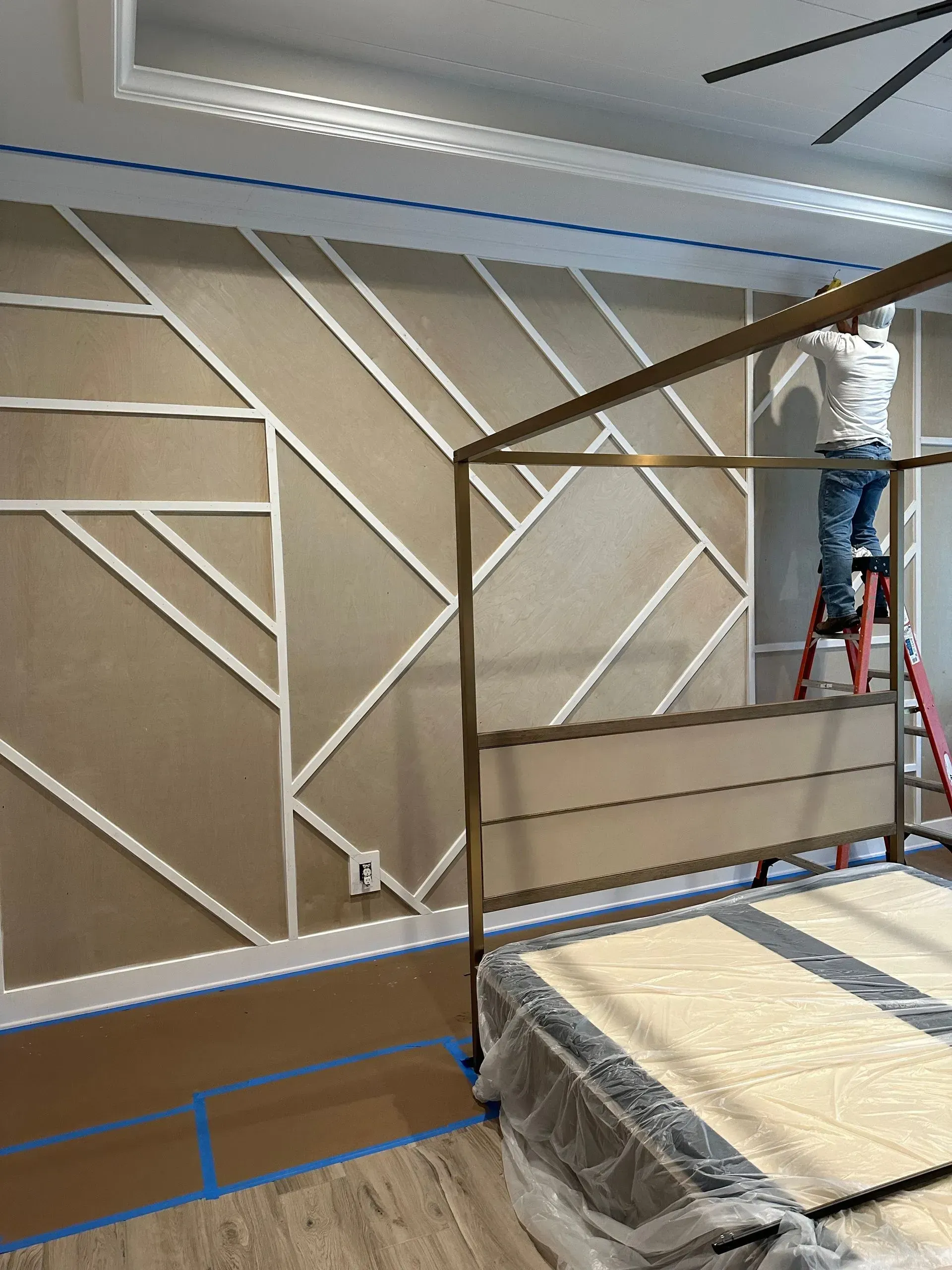 A person on a ladder installing trim on a geometric wall design in a bedroom with a canopy bed.