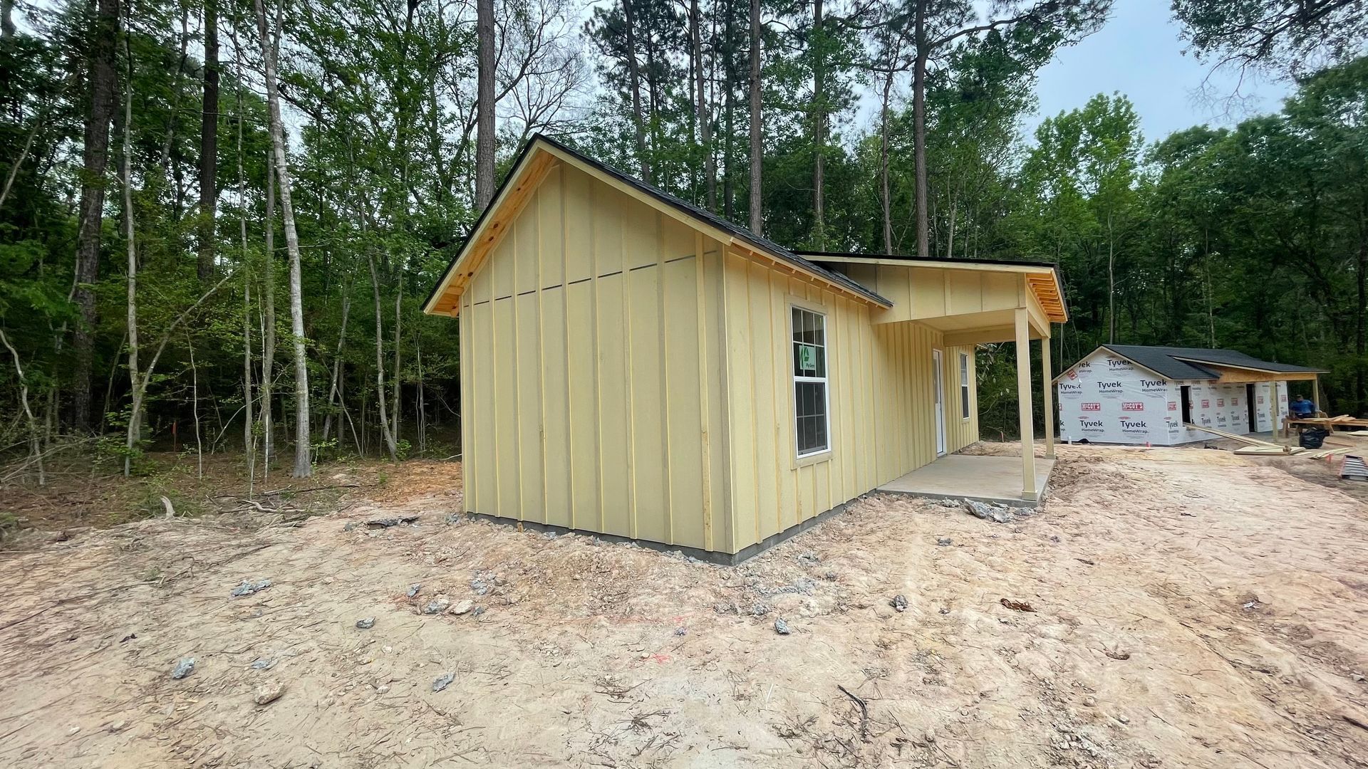 Two unfinished houses under construction in a wooded area, with light yellow siding on the front house and a small porch.