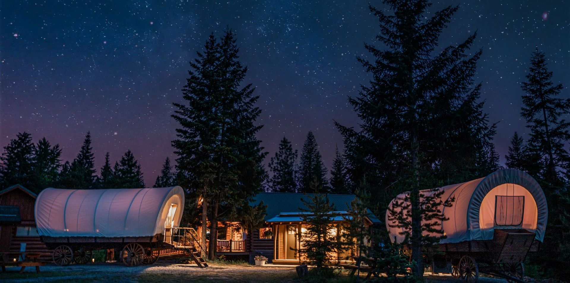 A covered wagon is parked in front of a house in the woods at night.