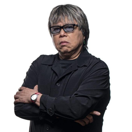 A man with short grey hair and black-framed glasses, wearing a black outfit, standing with arms crossed and a serious expression against a plain light background.