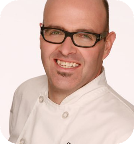 
A smiling man with glasses and a small goatee, wearing a white chef's jacket, facing slightly to the side against a light background.