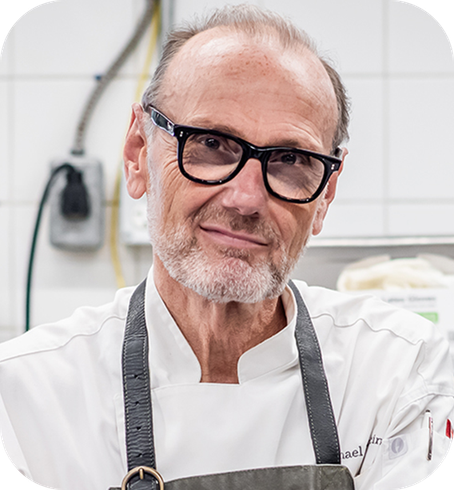 
Portrait of a slightly smiling man with a grey beard, wearing black glasses, a white chef's coat, and a dark apron, in a professional kitchen setting.
