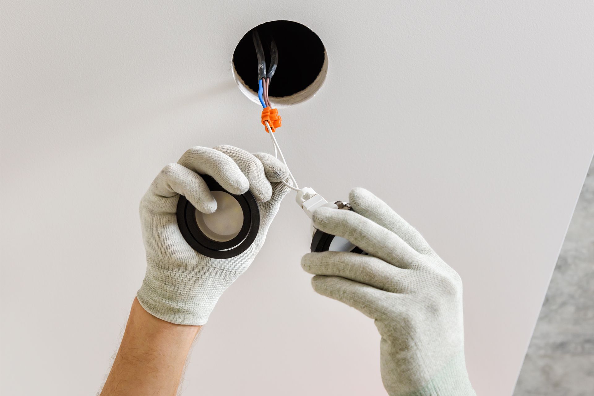 Gloved hands installing a wire connector into a ceiling junction box with exposed blue and orange wires.