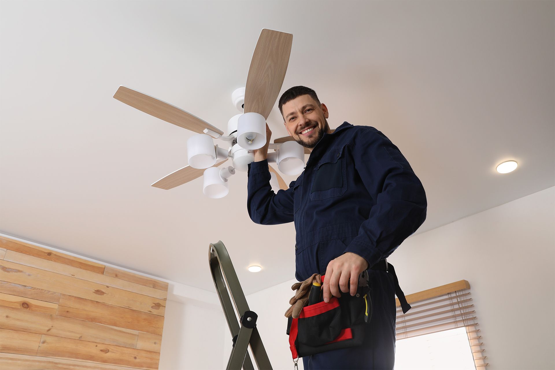 Person on a ladder installing a ceiling fan in a bright room.