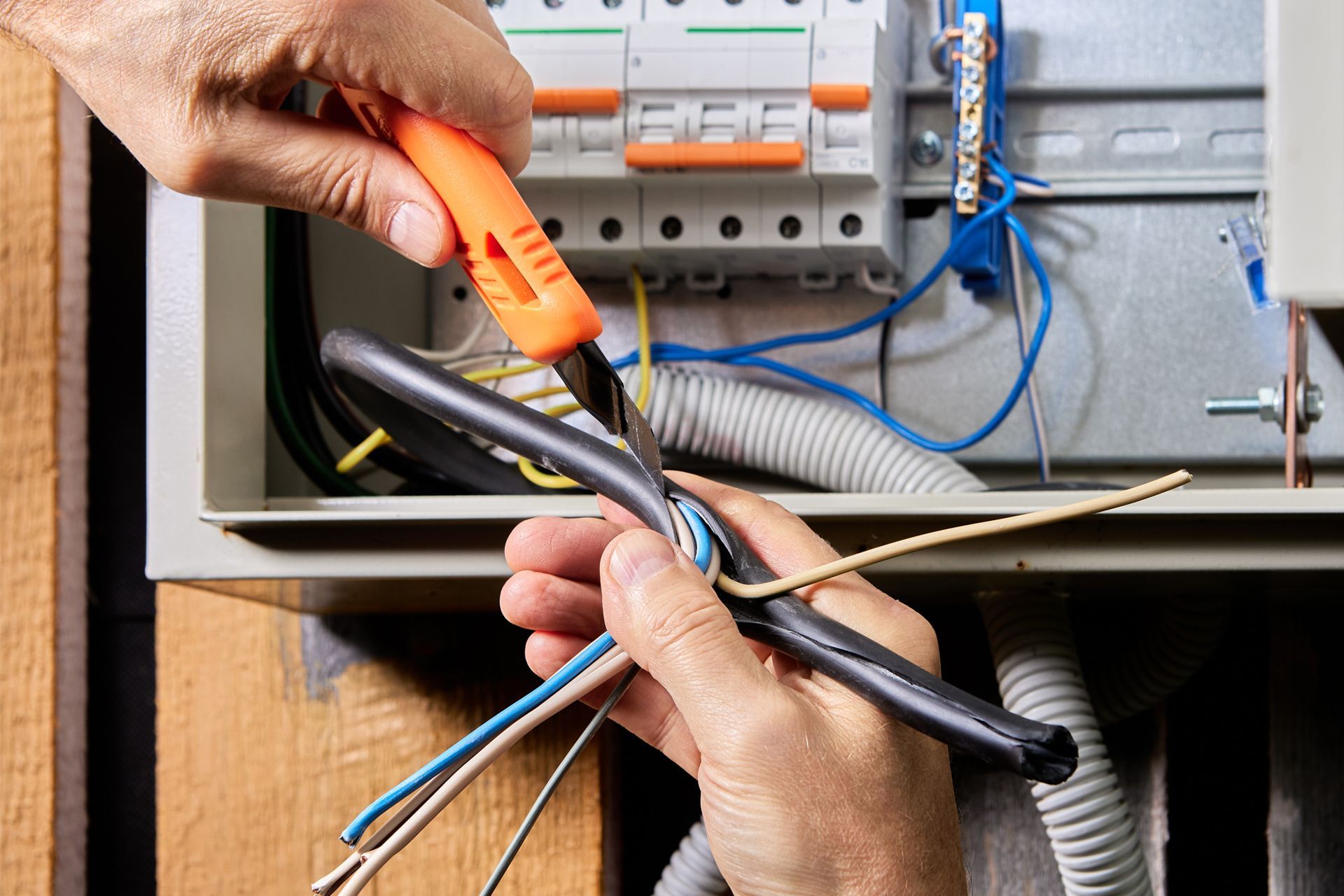 Hands using orange wire stripper to cut cables inside an open electrical panel.