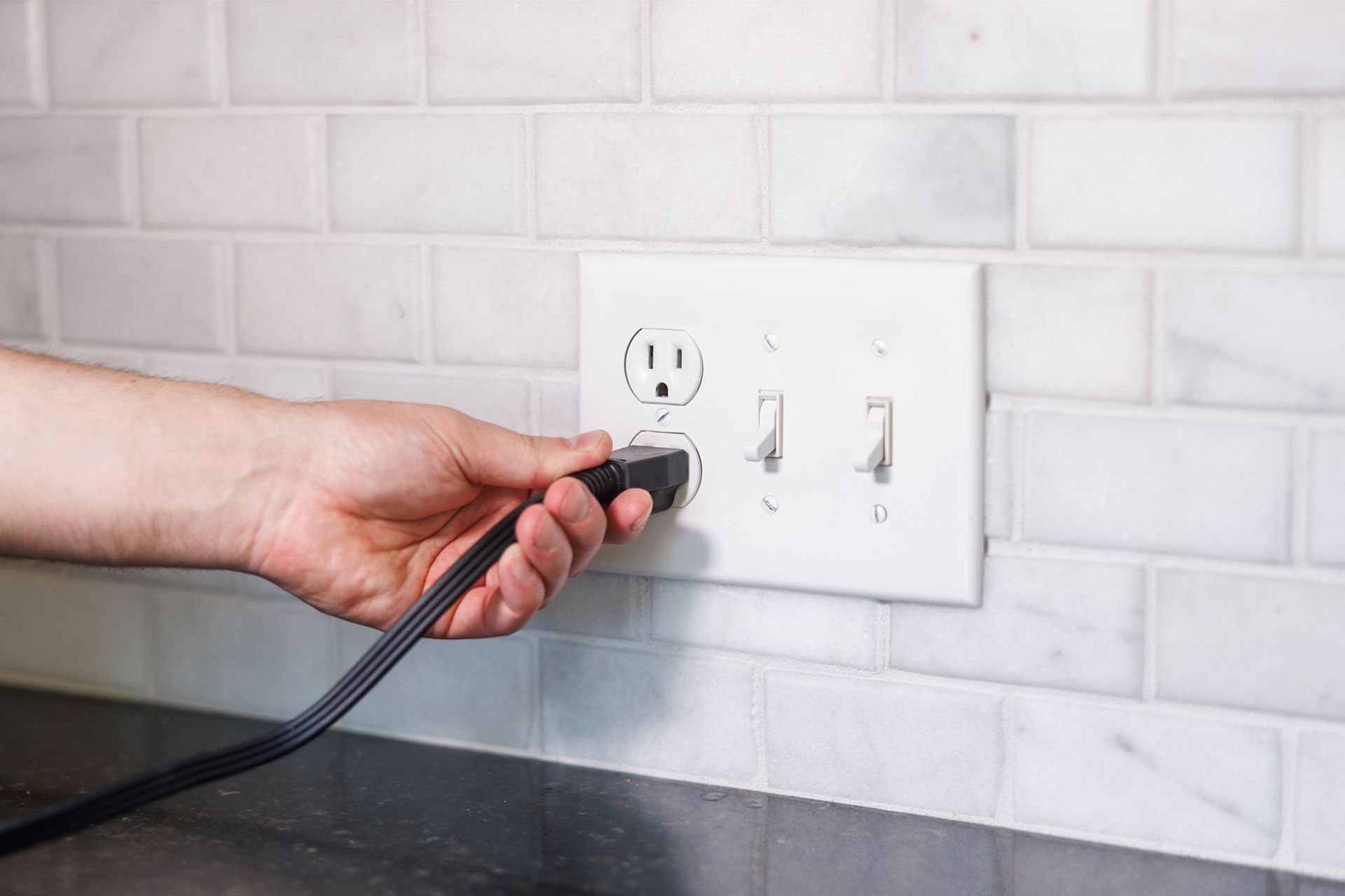 Hand plugging a black cord into a white wall outlet in a tiled kitchen.