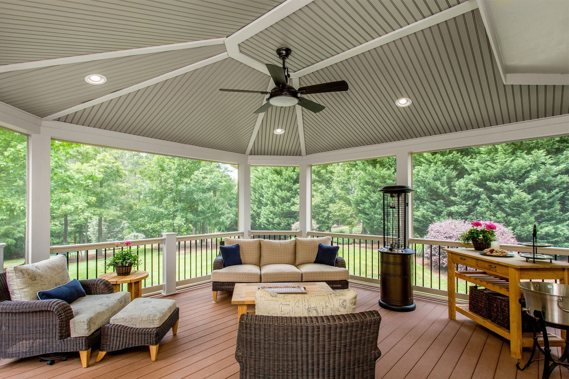 Covered outdoor patio with wicker seating, ceiling fan, wooden deck, and green wooded view.