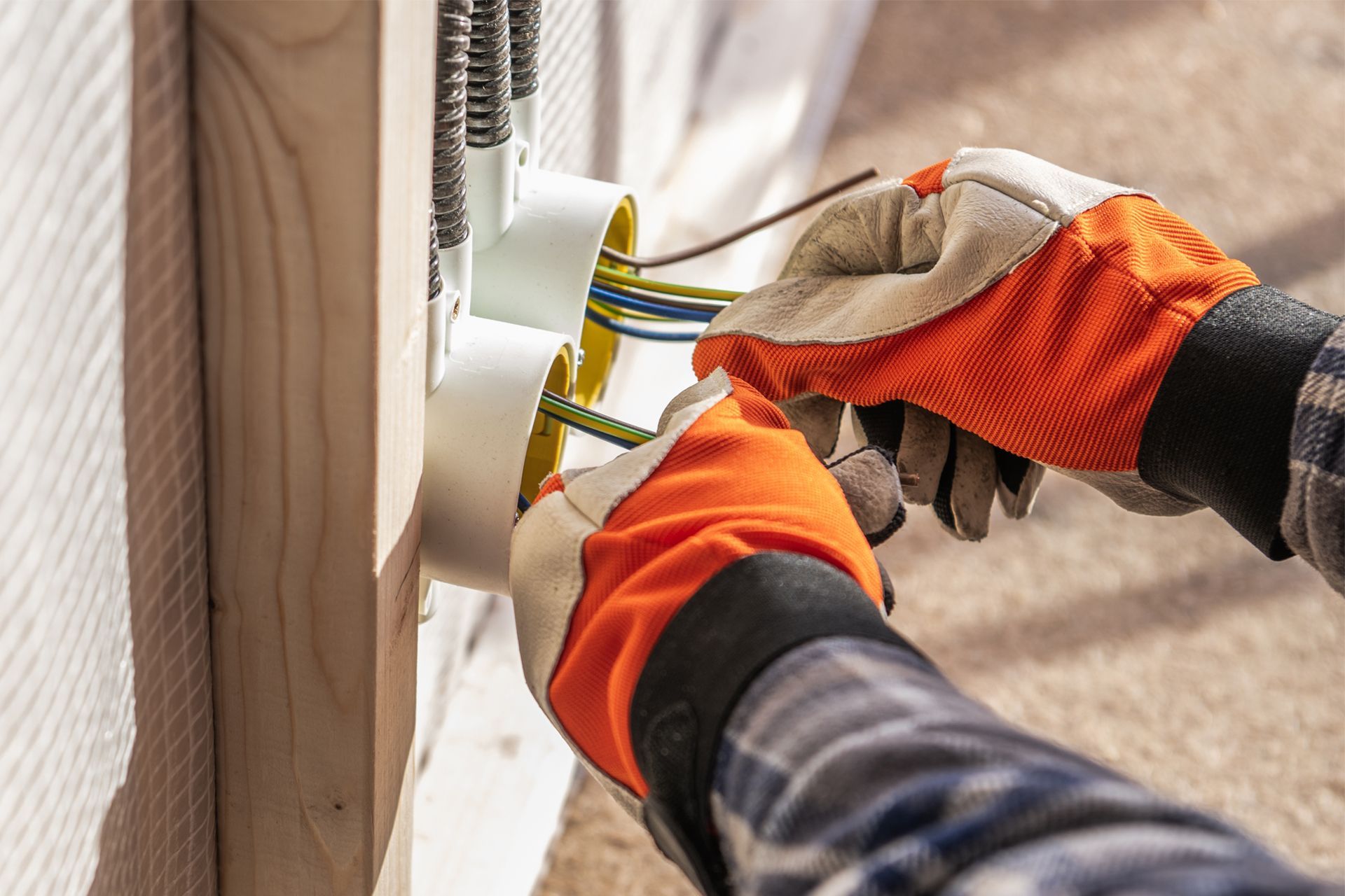 Gloved hands working on electrical wiring near a wall outlet, with orange and gray work gloves.