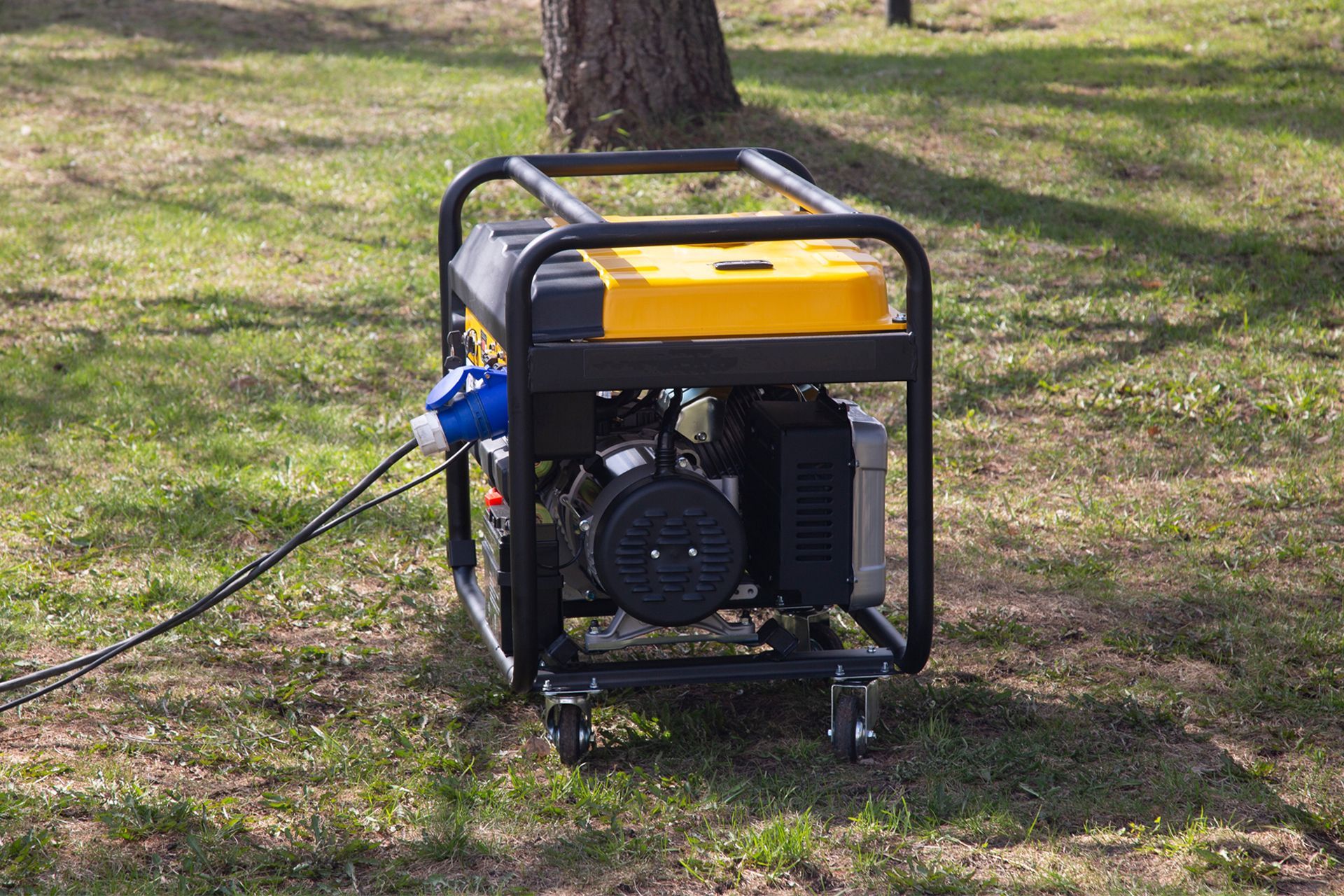Portable generator in a black frame with yellow tank on grass near a tree, plugged into an extension cord.