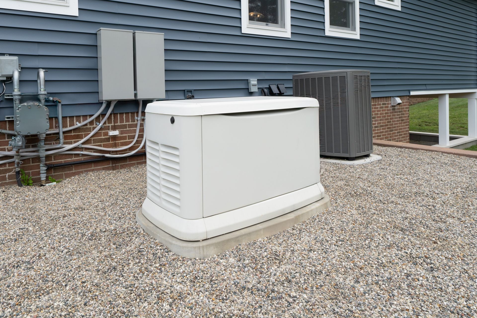 White outdoor generator on gravel beside a blue house and HVAC unit.
