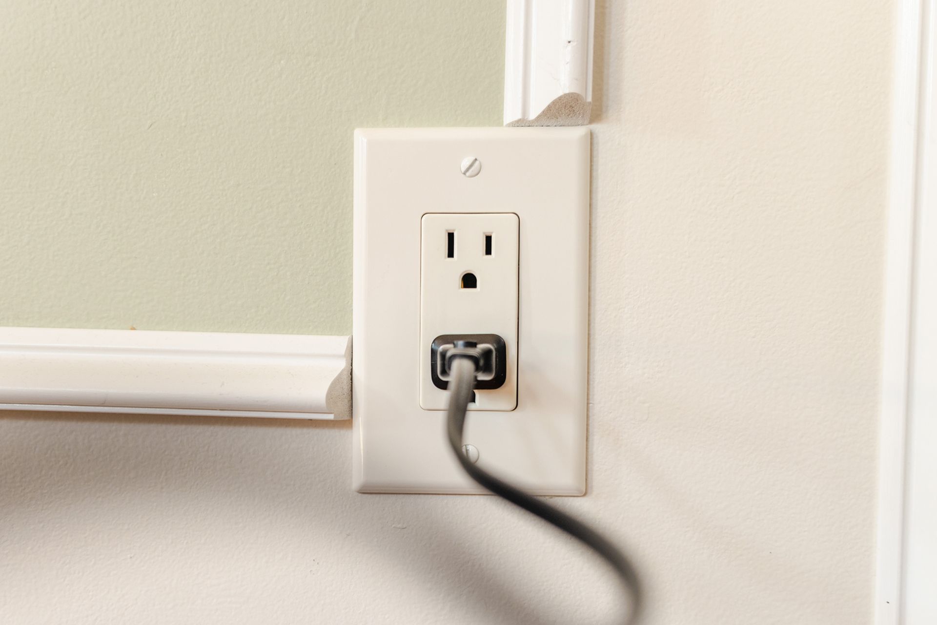 White wall outlet with a black plug inserted beside a light-colored chair rail.