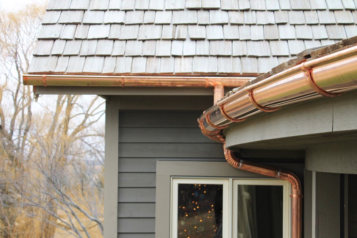Copper gutters on a gray house roof corner above a window, with trees in the background
