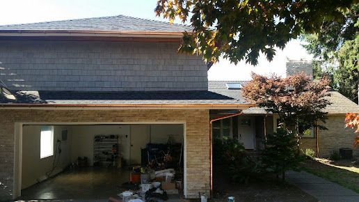 Brick house with open garage, dark shingled roof, and a shaded driveway with trees nearby