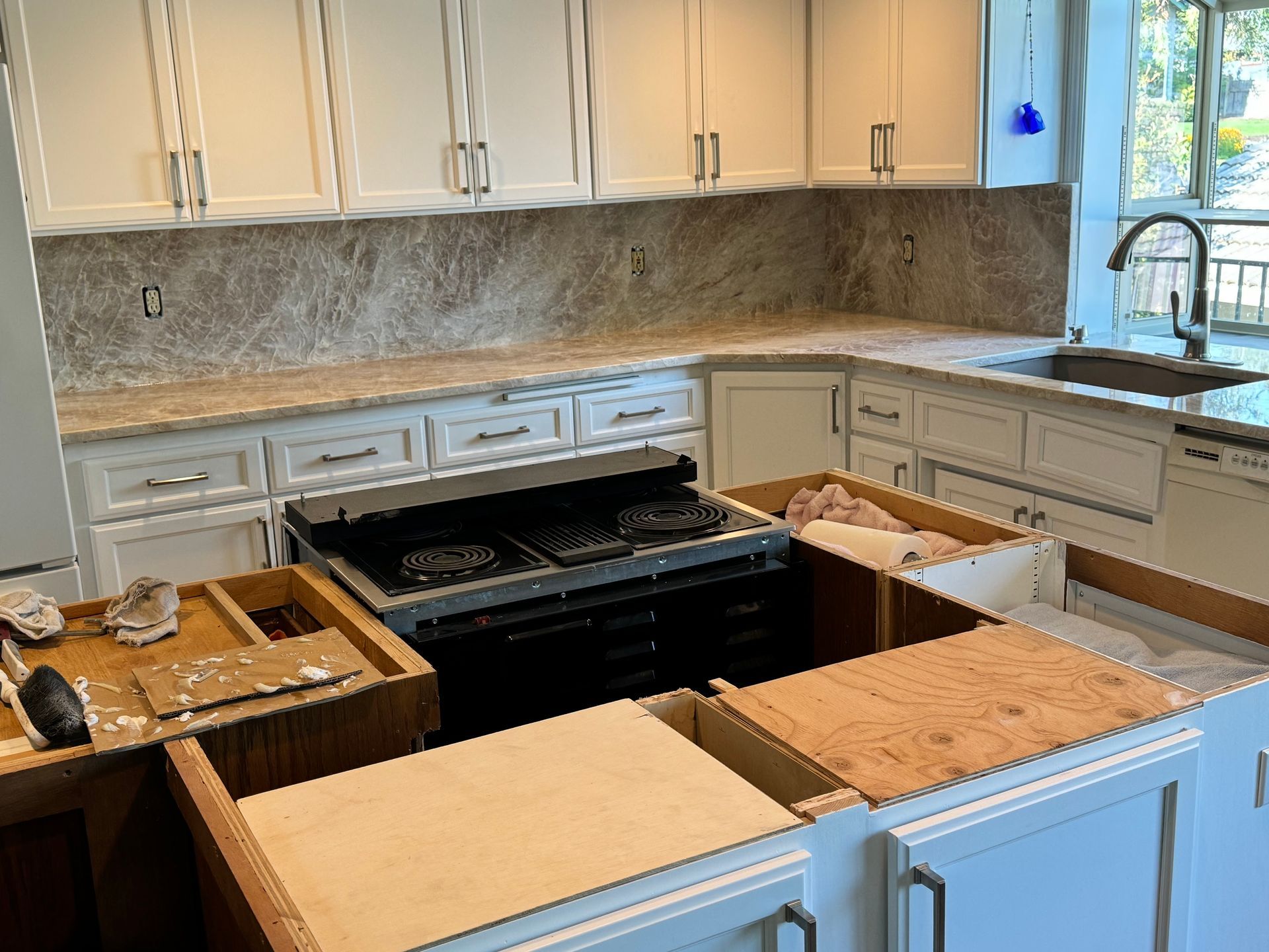 Kitchen under renovation with white cabinets, black stove, and countertops removed exposing plywood and open boxes