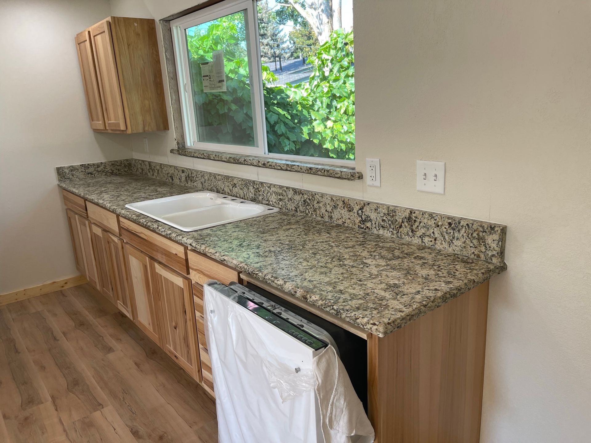 Empty kitchen counter with granite top, sink, upper cabinet, and window overlooking greenery