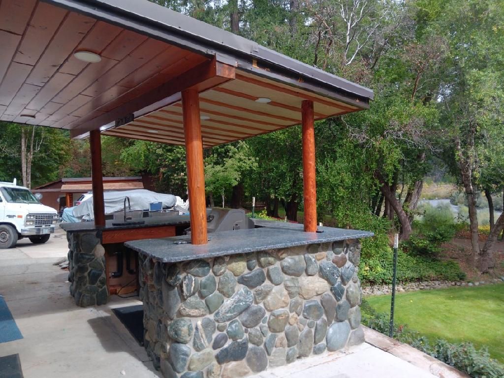 Stone outdoor shelter with wooden roof and posts beside a driveway and greenery