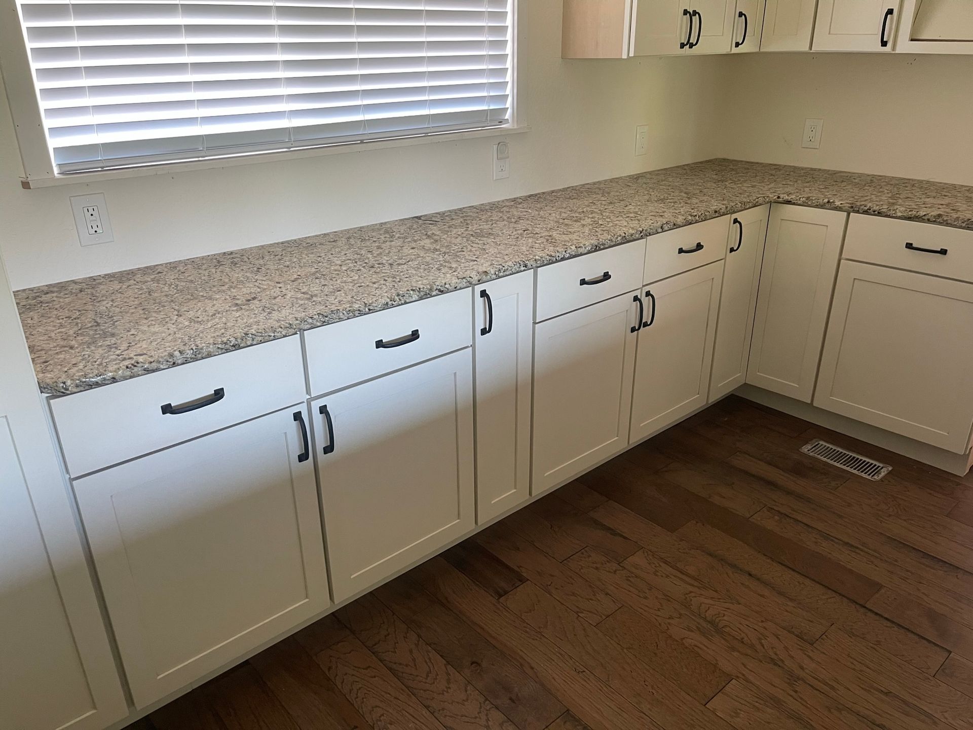 Corner kitchen with white cabinets, speckled countertops, window blinds, and wood-look flooring