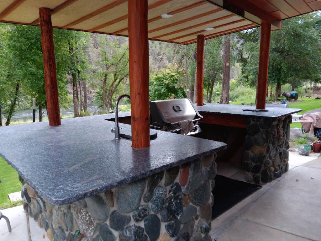Covered outdoor barbecue shelter with stone counters, grill, and a wooden roof in a wooded park.