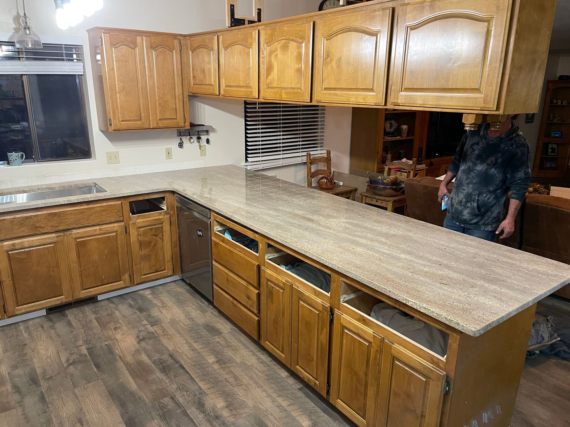 Empty kitchen with wood cabinets, granite countertops, and dark wood flooring; a person stands at the right edge.