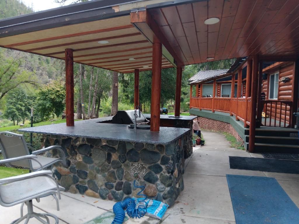 Covered stone patio with outdoor sink, red posts, metal chairs, and a log cabin walkway surrounded by greenery