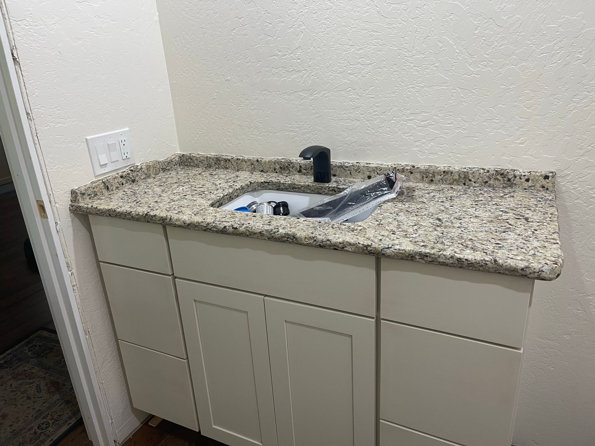 Bathroom vanity with beige cabinets, speckled countertop, and sink with black faucet against a light wall