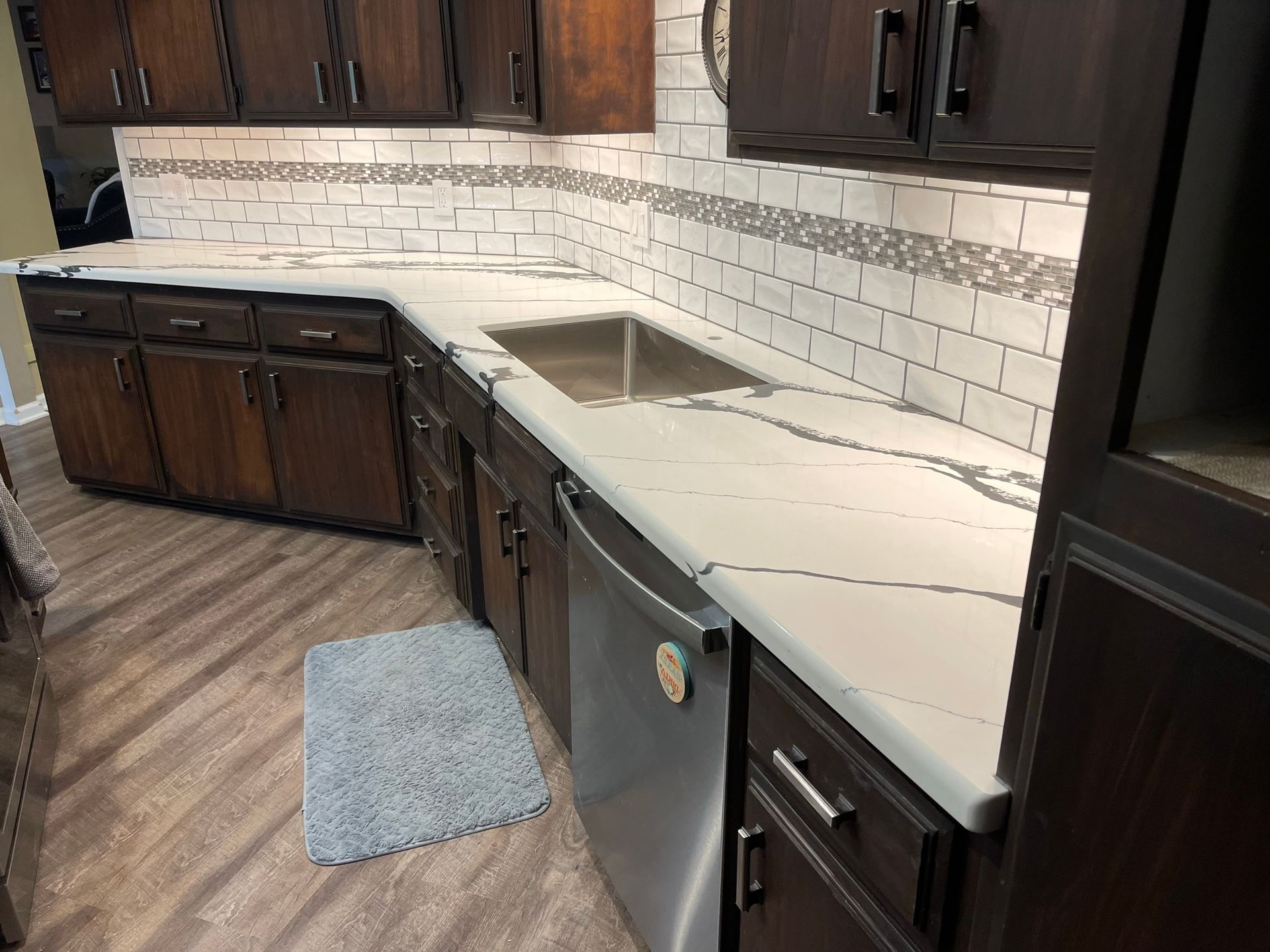 Modern kitchen with dark wood cabinets, light countertops, sink, dishwasher, and a blue rug.