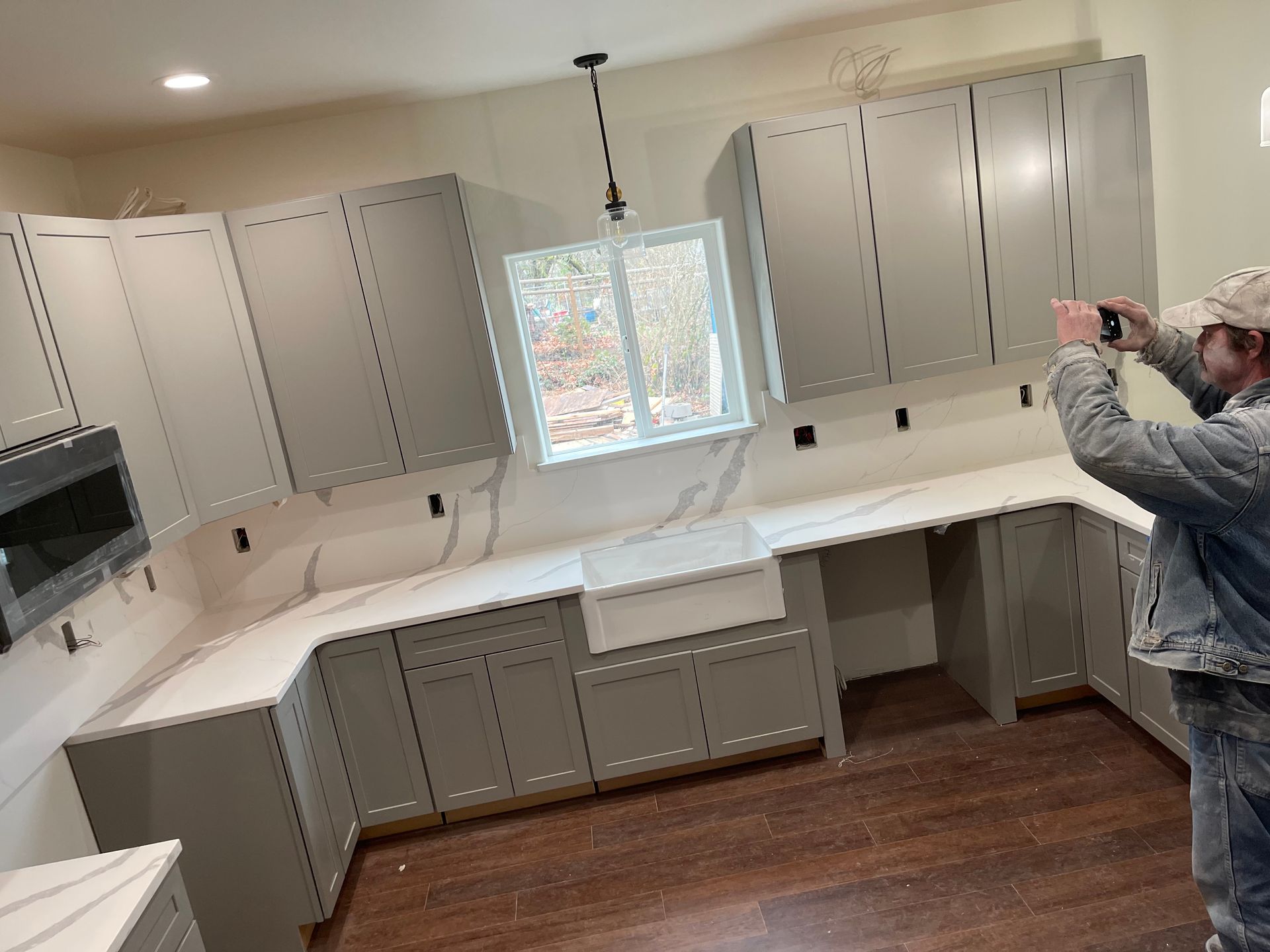 Kitchen under renovation with gray cabinets, white counters, and a person photographing the space