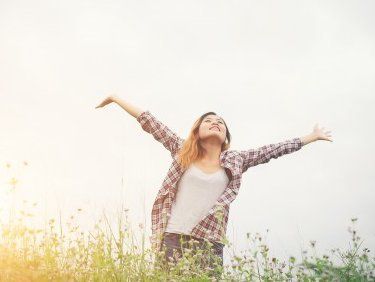 Woman with arms raised in a field of flowers, smiling, sunny sky.
