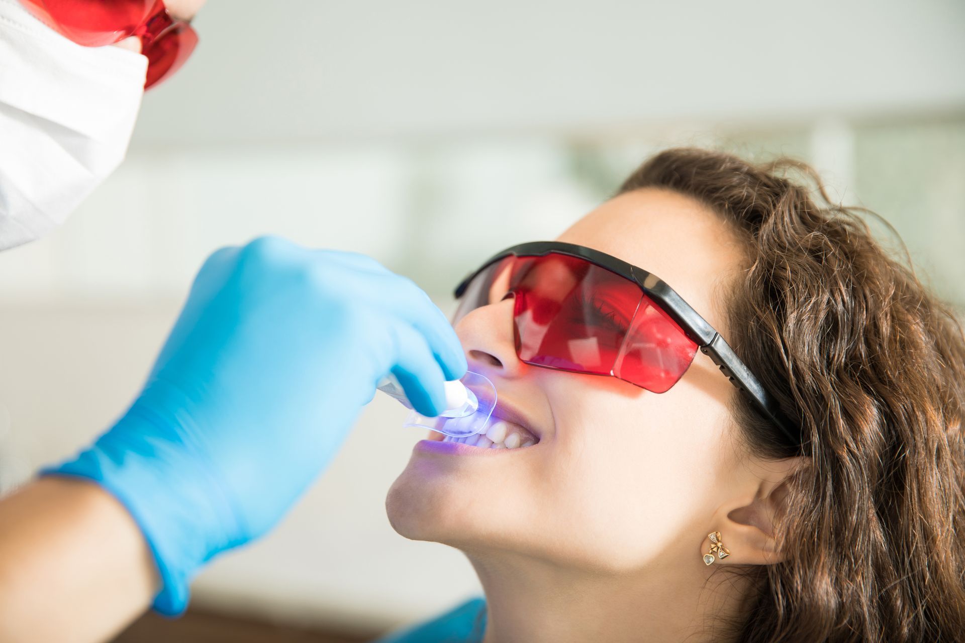 Dentist wearing blue gloves applies a light to a patient’s teeth, with red protective glasses on.