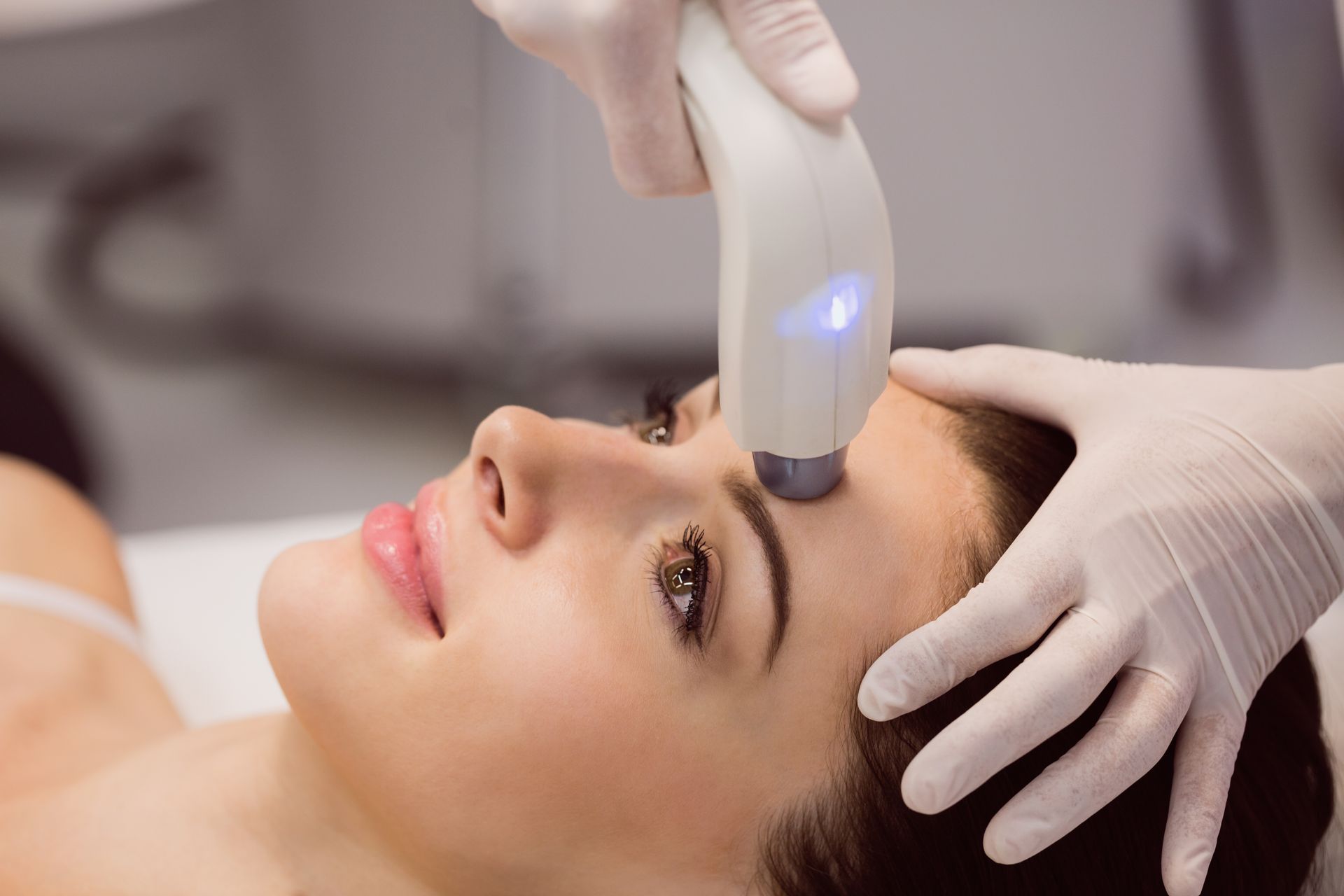 Woman receiving facial treatment with a handheld device on her forehead. Hands in gloves, indoor setting.