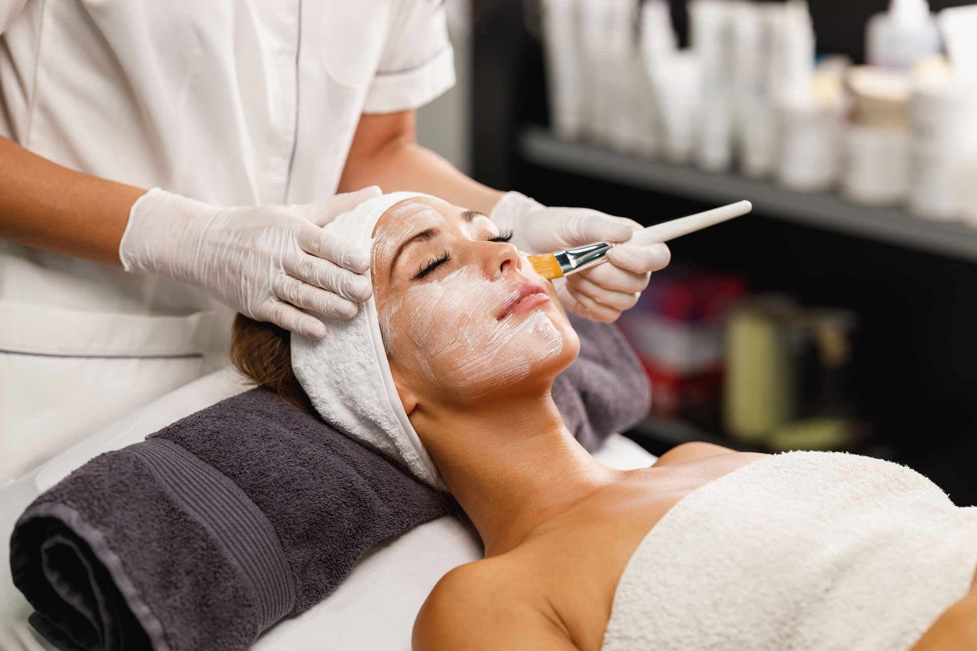 Facial treatment in spa, woman lying under towel as esthetician applies cream with a brush