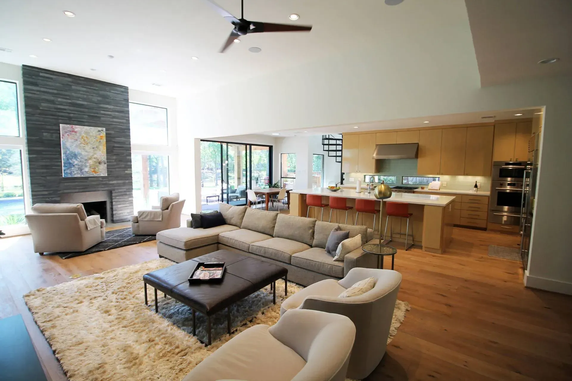 Modern open-plan living room with gray sectional, beige chairs, coffee table, and adjacent kitchen/bar area