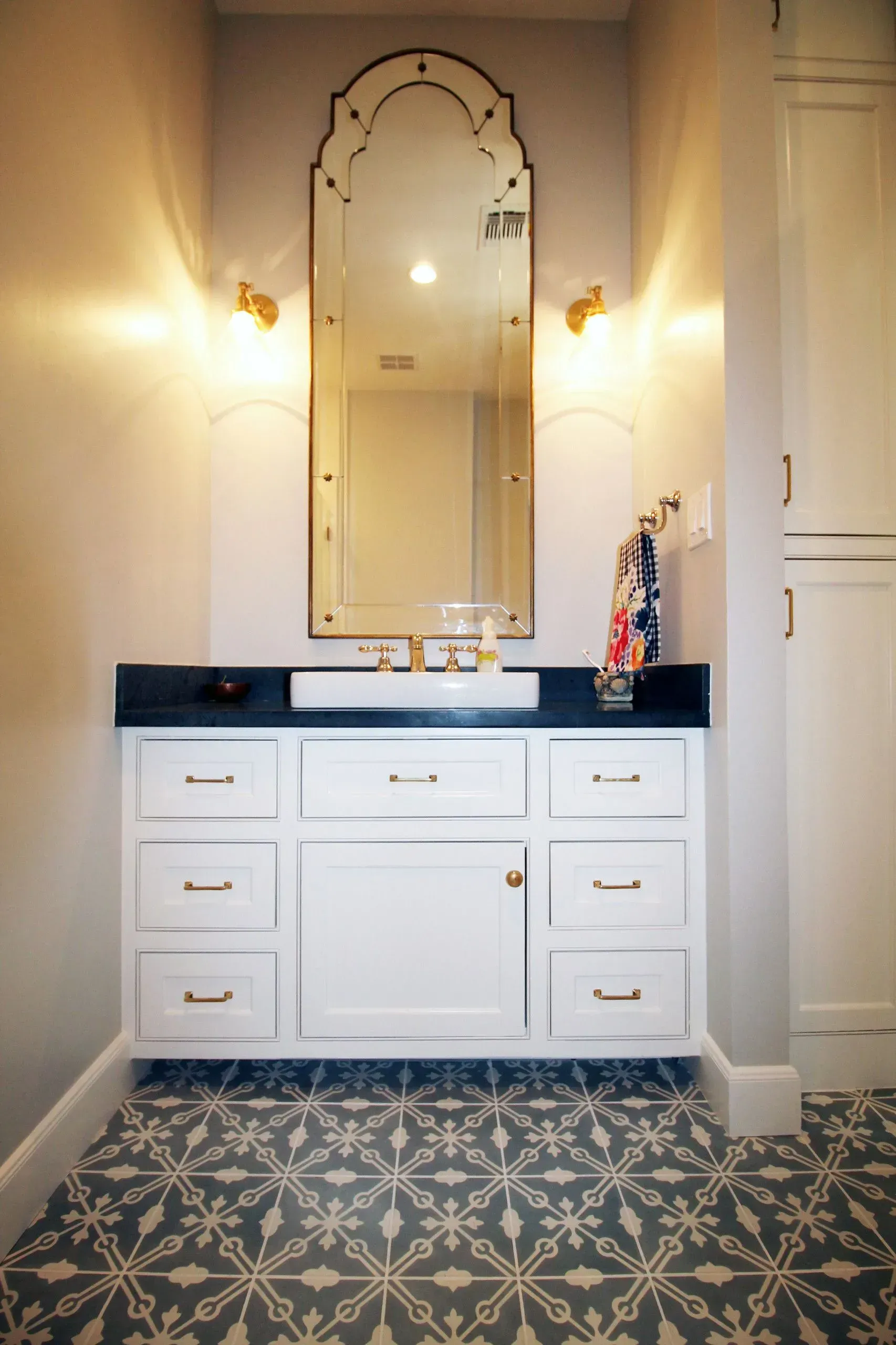 White bathroom vanity with ornate mirror, wall sconces, and patterned tile floor
