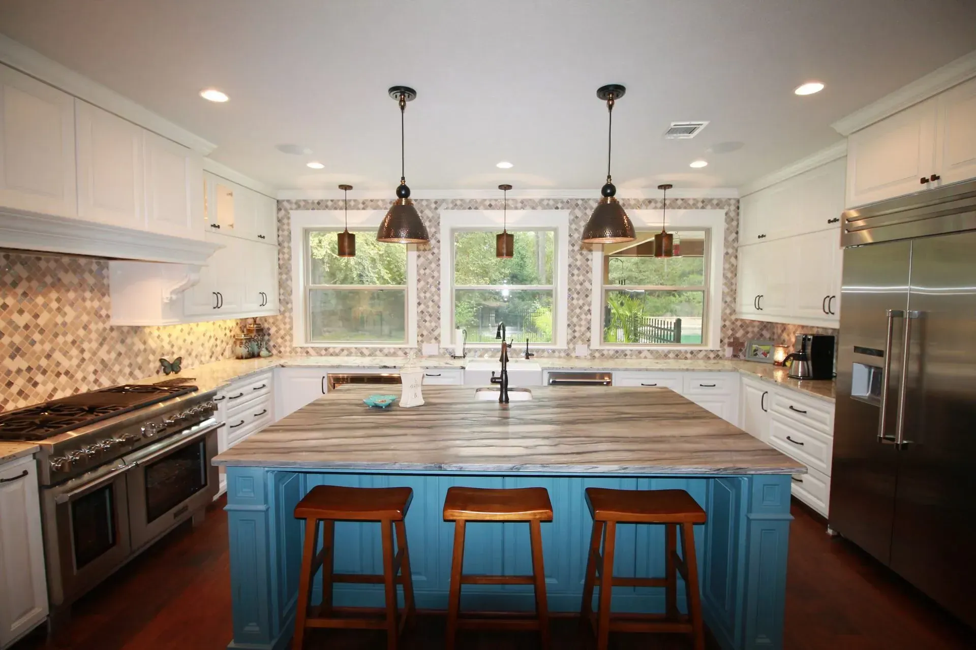 Bright modern kitchen with blue island, wooden stools, white cabinets, stainless appliances, and large windows