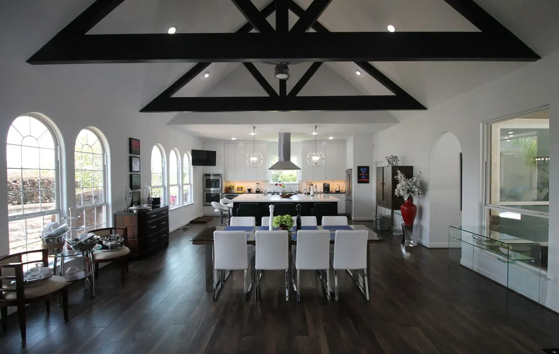 Modern open kitchen and dining area with high ceilings, dark floors, white chairs, and large windows