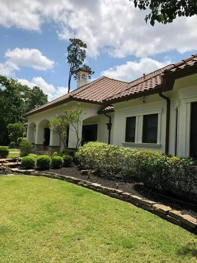 Spanish-style house with red-tile roof, white stucco walls, and a manicured green lawn under a partly cloudy sky