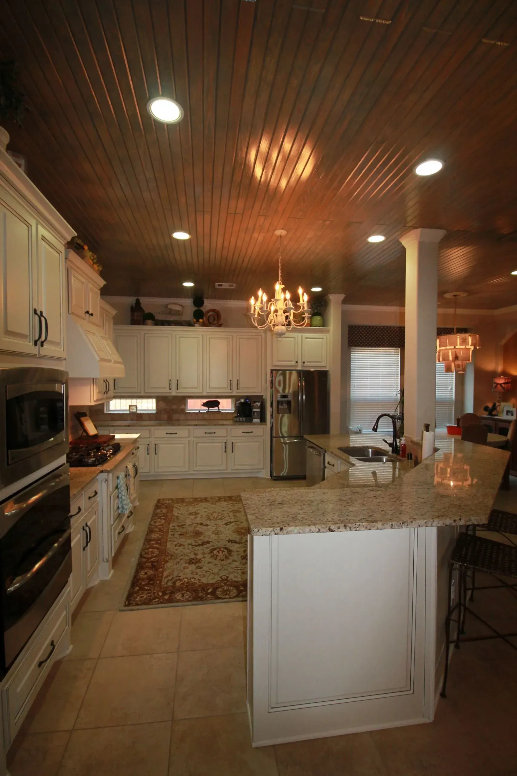 Spacious kitchen with white cabinets, granite island, pendant lights, and chandelier under a wood-paneled ceiling